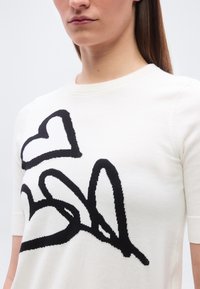 Γυναίκα που φοράει λευκό κοντομάνικο τοπ με μεγάλο μαύρο αφηρημένο σχέδιο καρδιάς και γραμμής στο στήθος.