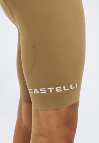 Κοντινό πλάνο σε μπεζ ποδηλατικό σορτς στο μηρό, με λευκό λογότυπο "CASTELLI" και λεπτομέρειες ραφής με επίπεδη ραφή.