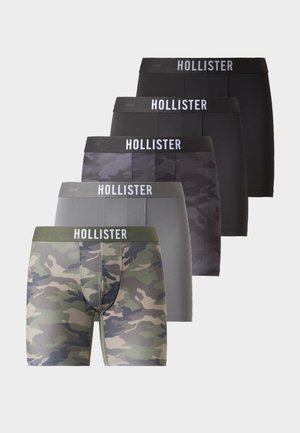 Mężczyzna bez koszulki stoi boso, ubrany w ciemnoszare bokserki moro z paskiem "Hollister" na jasnym, jednolitym tle.