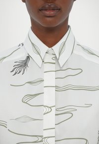 Cămașă albă cu nasturi, decorată cu modele abstracte verzi și accente negre pe guler și plastron. Textură de țesătură netedă.