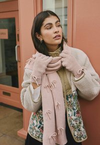 Sciarpa rosa lavorata a maglia con dettagli a fiocco, giacca in pile beige con motivo floreale e guanti coordinati. Tessure e colori sovrapposti.