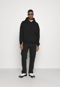 Sort bomulds-hættetrøje med snoretræk, frontlomme, parret med sorte jeans og hvide sneakers med sort sål. Klare, tonede solbriller.
