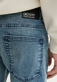 Jeans de mezclilla azul claro con un detalle en el bolsillo trasero, que presenta una etiqueta negra que dice "DENIM TOM TAILOR" y acentos de costura.