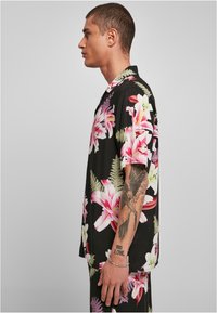 Cămașă neagră cu mânecă scurtă, cu un model floral roz și alb, cu guler, croială lejeră și tatuaj vizibil pe braț.