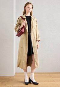 Nainen, jolla on yllään beige trenchcoat mustan mekon päällä, valkoiset sukat, mustat korkokengät ja punainen käsilaukku, seisoo puulattialla yksinkertaista seinää vasten.