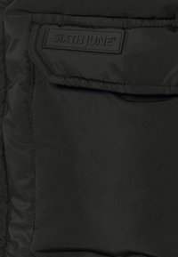 Zbliżenie czarnej tkaniny pokazujące kieszeń z klapką i naszywkę z napisem "SIXTH JUNE" w lewym górnym rogu.