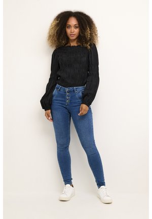 Top noir à manches longues smocké associé à un jean skinny taille haute bleu et des baskets blanches. Tissu texturé et détail de devant à cinq boutons.