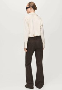 Camicia beige chiaro con bottoni, in tessuto liscio, abbinata a pantaloni marroni a zampa d'elefante in lana testurizzata. Senza motivi o elementi visibili.