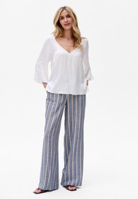 Weiße Bluse mit langen Ärmeln und V-Ausschnitt, kombiniert mit blauen und gelben gestreiften, weit geschnittenen Hosen. Glatter Stoff mit lockerer Passform.
