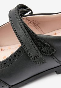 Schwarzer Lederschuh mit einem Klettverschluss, ausgestattet mit perforierten Details und einer weichen, rosa Sternenmuster-Innensohle. Flache Sohle.