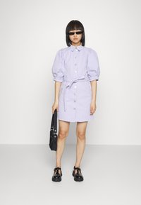 Vestido lilás de botões com mangas bufantes, cintura ajustada e cinto coordenado. Combinado com acessórios pretos e sapatos chunkys.