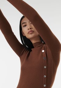 Pull en ribbed marron avec un col haut, comportant des boutons argentés le long du côté gauche et un design ajusté. Tissu texturé.