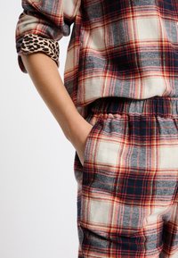 Blusa de cuadros rojos y marinos con mangas enrolladas, combinada con pantalones de cuadros con cintura elástica. Ambos cuentan con una textura suave y tejida, con un acento de estampado de leopardo.