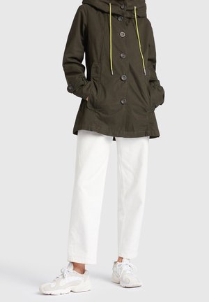 Kobieta w ciemnozielonej kurtce z kapturem z żółtymi troczkami, białe spodnie z szerokimi nogawkami i białe trampki, stojąca z rękami w kieszeniach.