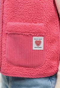 Veste polaire rose avec poche côtelée et patch affichant un design cœur et un texte disant "Wild at heart, free in spirit."
