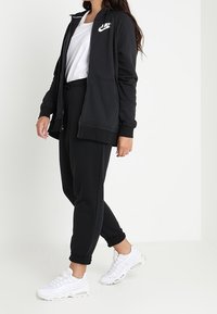 Svart zip-up hoodie med vitt Nike-logotyp, parat med svarta joggerbyxor och vita sportskor, mot en neutral bakgrund.