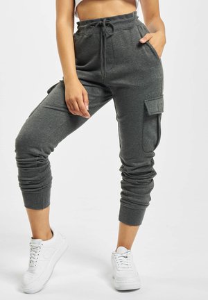 Kobieta ubrana w ciemnoszare joggery typu cargo z troczkiem w pasie i białe trampki, stoi z jedną nogą zgiętą i ręką w kieszeni.