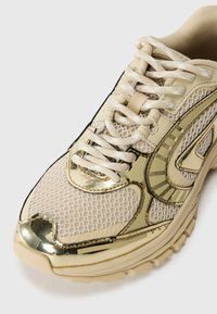 Beige treningssko med mesh-stoff og gullmetalliske aksenter. Har teksturert overflate, rund tå og tradisjonell snøredesign.