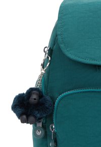 Zaino in tessuto turchese con chiusura a pattina. Presenta una superficie testurizzata e un ciondolo a forma di gorilla con un corpo peloso di un blu scuro.