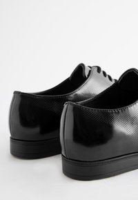 Black patent leather shoes with a smooth texture, rounded design, and perforated detailing on the back. Rubber soles with a subtle tread pattern.