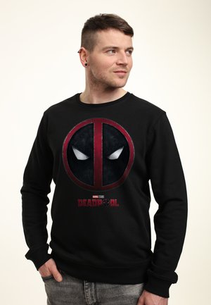 Man met kort haar en oorbellen die een zwarte Deadpool-trui draagt, staat met één hand in zijn zak en kijkt naar rechts tegen een effen achtergrond.