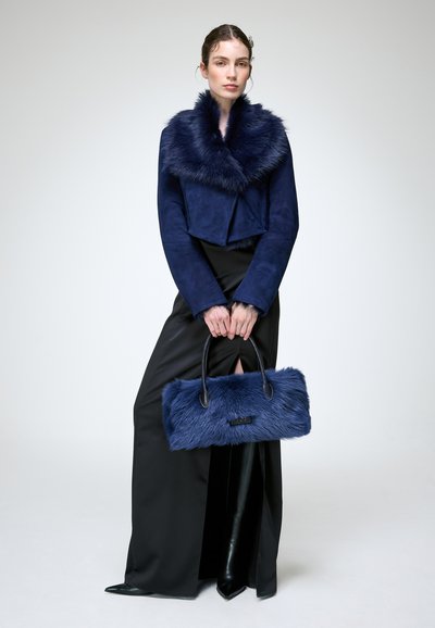 Femme portant une veste à col en fourrure bleu marine et une jupe fendue noire, tenant un sac à main assorti en fourrure bleu marine avec des poignées noires.