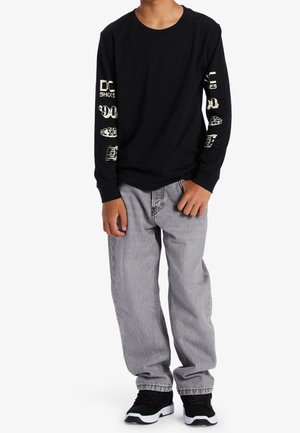 Młoda osoba ubrana w czarną bluzę z długim rękawem z białym napisem na rękawach, luźne szare dżinsy oraz czarne trampki z białą podeszwą, stojąca na białym tle.