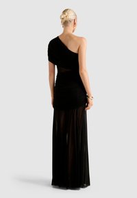 Sieviete ar blondiem matiem augstā bulciņā, tērpusies melnā vakarkleitā ar vienu plecu, caurspīdīgu svārku paneļu, valkā zelta auskarus un aproci, stāv ar muguru pret skatītāju.