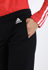 Gros plan sur une main dans la poche d'un pantalon noir Adidas porté avec un haut à manches longues rouge doté de rayures blanches sur la manche.