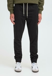 Black, tapered sweatpants with elastic cuffs and a drawstring waistband, paired with white and gray athletic shoes.