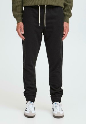 Black, tapered sweatpants with elastic cuffs and a drawstring waistband, paired with white and gray athletic shoes.