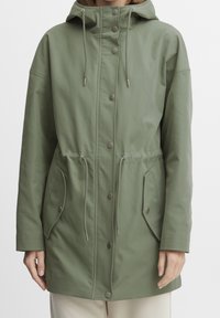 Giacca parka verde realizzata in un morbido tessuto testurizzato. Presenta un cappuccio con cordino, bottoni a pressione e due tasche frontali con toppe.