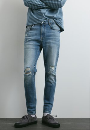 Personne debout, les bras croisés, portant un jean bleu déchiré, des baskets grises, des chaussettes blanches et une chemise à manches longues rayée bleue sur un fond uni.
