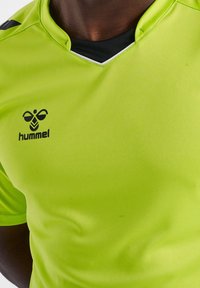 Jersey deportivo de un brillante color verde neón con el logotipo negro de hummel en el pecho y una camiseta negra visible en el cuello en forma de V.