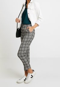 Kvinna klädd i vit jacka, mörkgrön blus, svartvit rutiga byxor, vita sneakers och bärande på en svart axelväska.