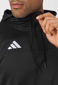 Schwarzer sportlicher Hoodie mit Reißverschluss am Kragen, strukturiertem Stoff und einem weißen Adidas-Logo auf der Brust. Die Kordeln sind am Kragen sichtbar.