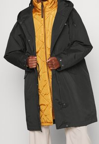 Veste extérieure noire avec capuche, associée à une couche intérieure matelassée jaune vif. Les deux comportent des fermetures éclair et des boutons-pression.