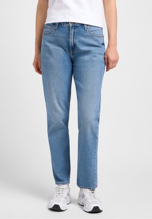 Persoon die lichtblauwe jeans met rechte pijpen, witte sneakers en een wit shirt draagt, staande tegen een effen achtergrond.