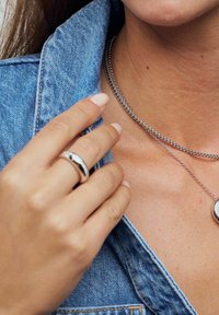 Silverring med texturerad design, buren på en hand som rör vid kragen av en blå denimjacka. Lager av silverkedjesmycken är också synliga.