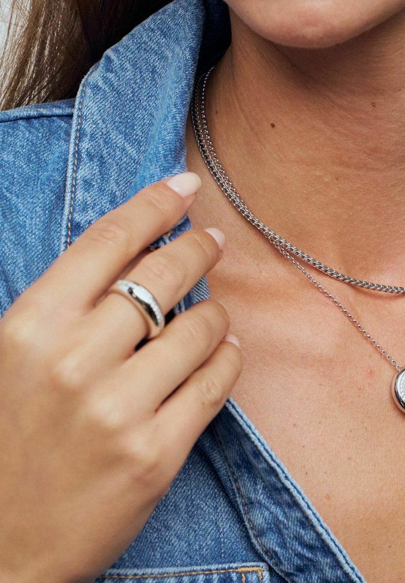 Silverring med texturerad design, buren på en hand som rör vid kragen av en blå denimjacka. Lager av silverkedjesmycken är också synliga.