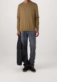 Homme portant une chemise marron à manches longues, un jean bleu délavé, des baskets noires, tenant un sac fourre-tout noir devant un fond uni.