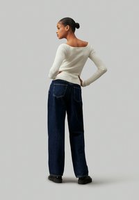 Top blanco de manga larga con cuello cuadrado, combinado con unos vaqueros de pierna ancha azul oscuro con bolsillos traseros y zapatos negros.