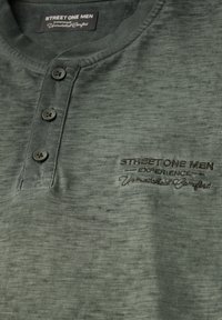 Tmavě zelené polo tričko s texturovaným povrchem, obsahující tři knoflíky a vyšívané logo na levé straně.