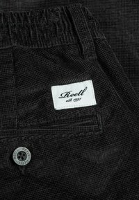 Nahaufnahme von einer schwarzen Cordhosen-Tasche mit einem weißen "Reell est. 1997" Etikett und einem schwarzen Knopf, der mit "Reell" graviert ist.