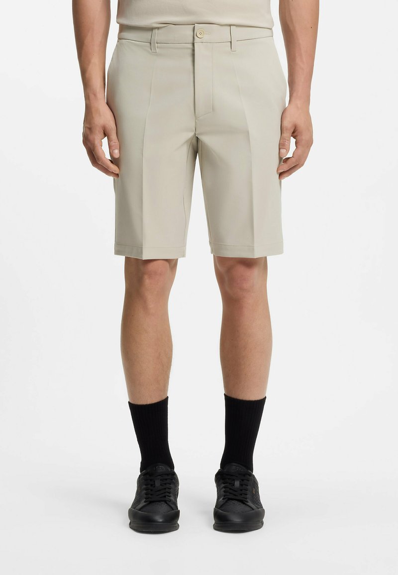 Mann trägt beige, knieumspielende, maßgeschneiderte Shorts, schwarze Socken und schwarze Sneakers, steht mit locker an den Seiten hängenden Armen.