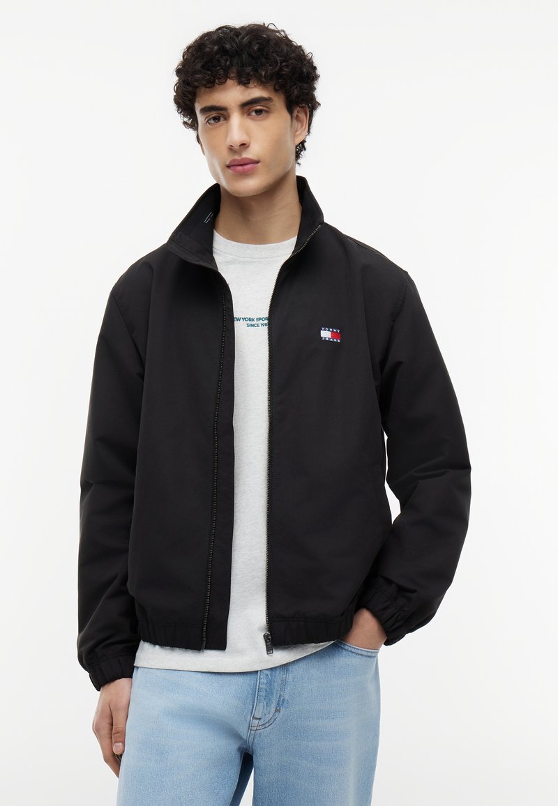 Joven con cabello rizado que lleva una chaqueta negra con cremallera sobre una camiseta blanca y jeans azul claro, posando frente a un fondo liso.