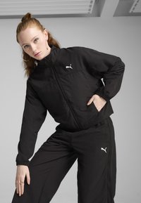 Črna trenerka in hlače iz lahkega materiala, z zadrgo spredaj, elastičnimi manšetami in belim logotipom Puma na prsih in stegnu.