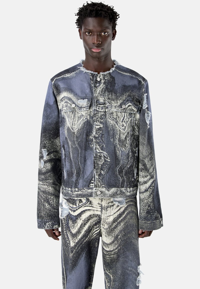 Jeune homme portant une veste en jean déchirée assortie à un pantalon avec des motifs abstraits noirs et blancs, debout devant un fond uni.