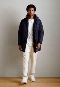 Cappotto imbottito blu navy con colletto ampio, abbinato a una maglietta bianca e pantaloni beige chiaro. Sneakers bianche completano il look.