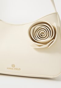 Bolso de mano de color crema, hecho de un material suave con un acento en forma de rosa. Presenta un logo de la marca en relieve y un diseño curvado y minimalista.